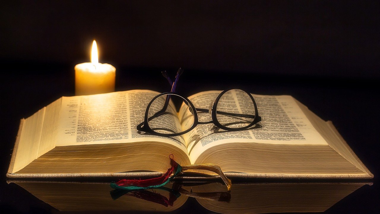 An open Bible with reading glasses placed on top of the open page, and a canddle lit up next to the Bible