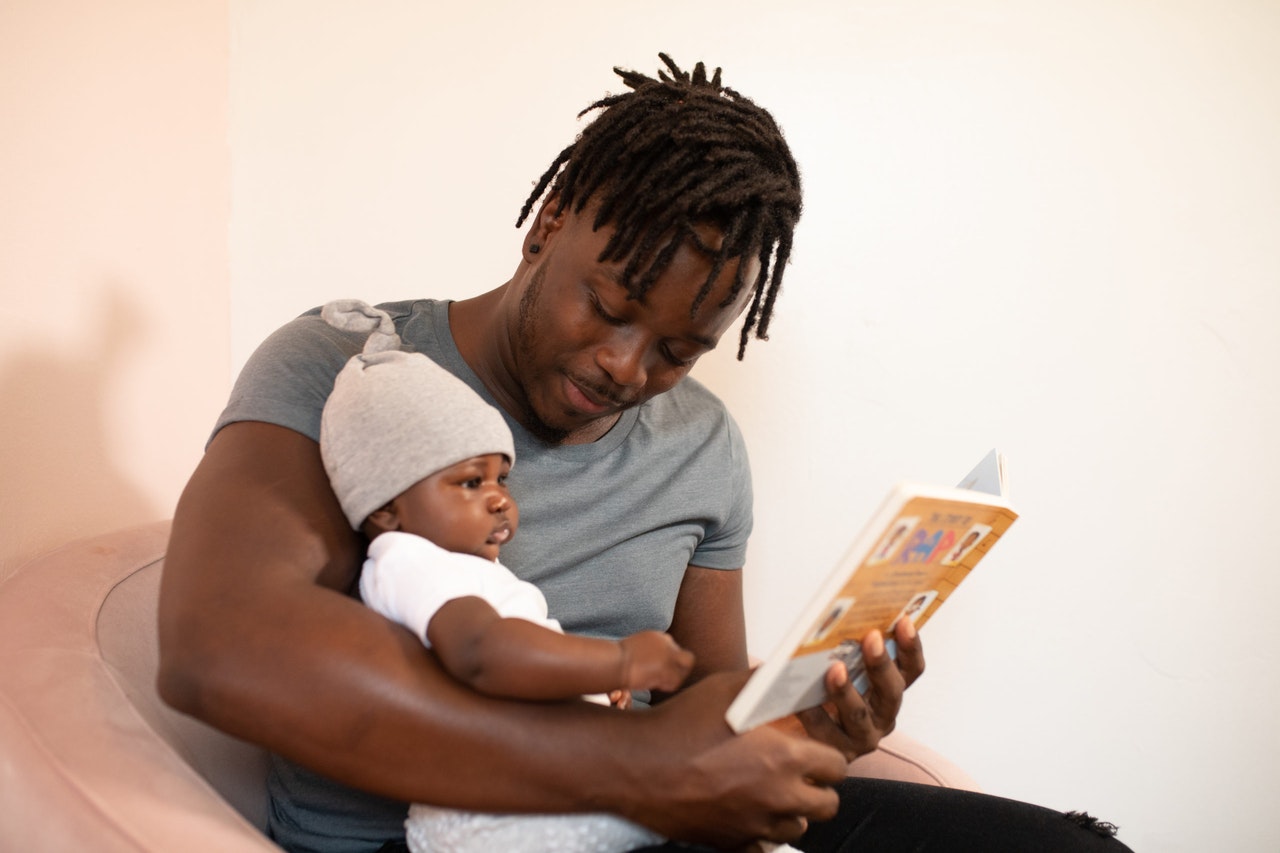 Man in grey holding a baby in white onsie and reading out of a book