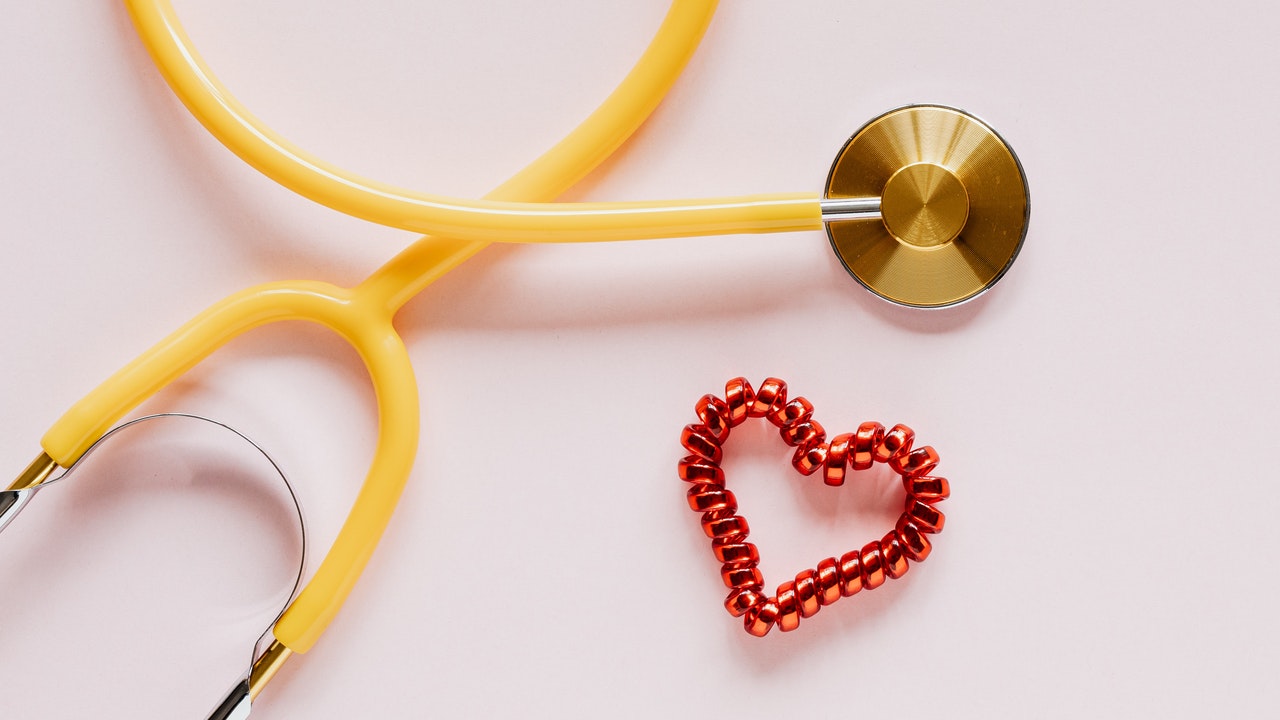 Yellow Stethoscope near red decorative coil tie in heart shape on pink surface.