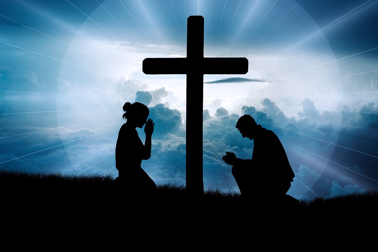 Silhuette of two people kneeling in humility befor the cross with heavenly light shinning on them.
