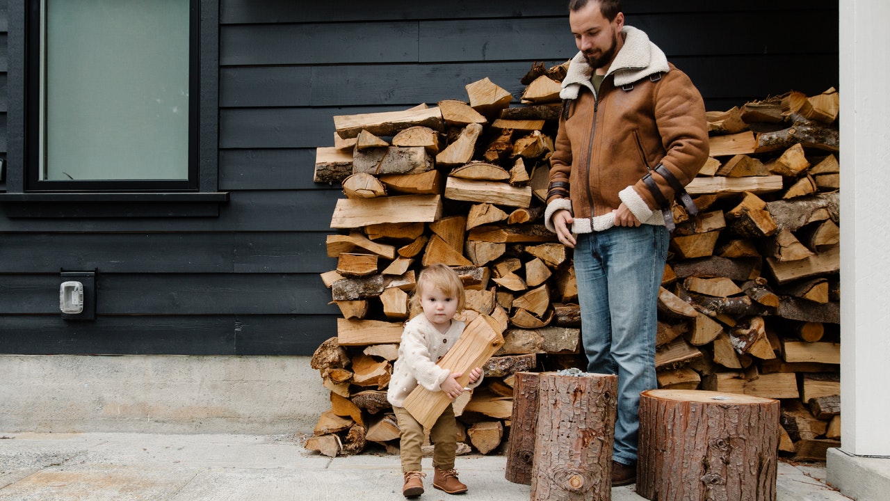A Child carrying a heavy piece of log with an adult watching to assist.