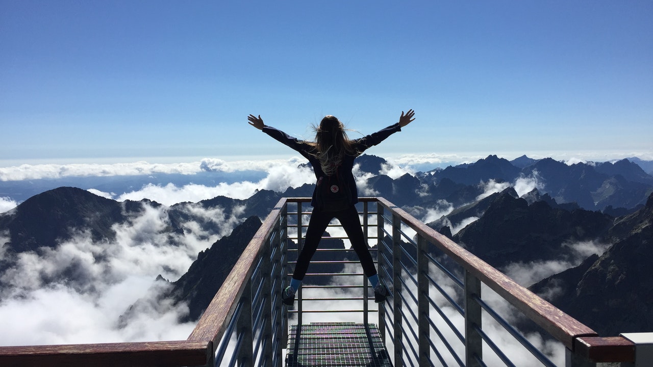 A Person standing on hand rails with arm spread apart facing mountains and clouds.