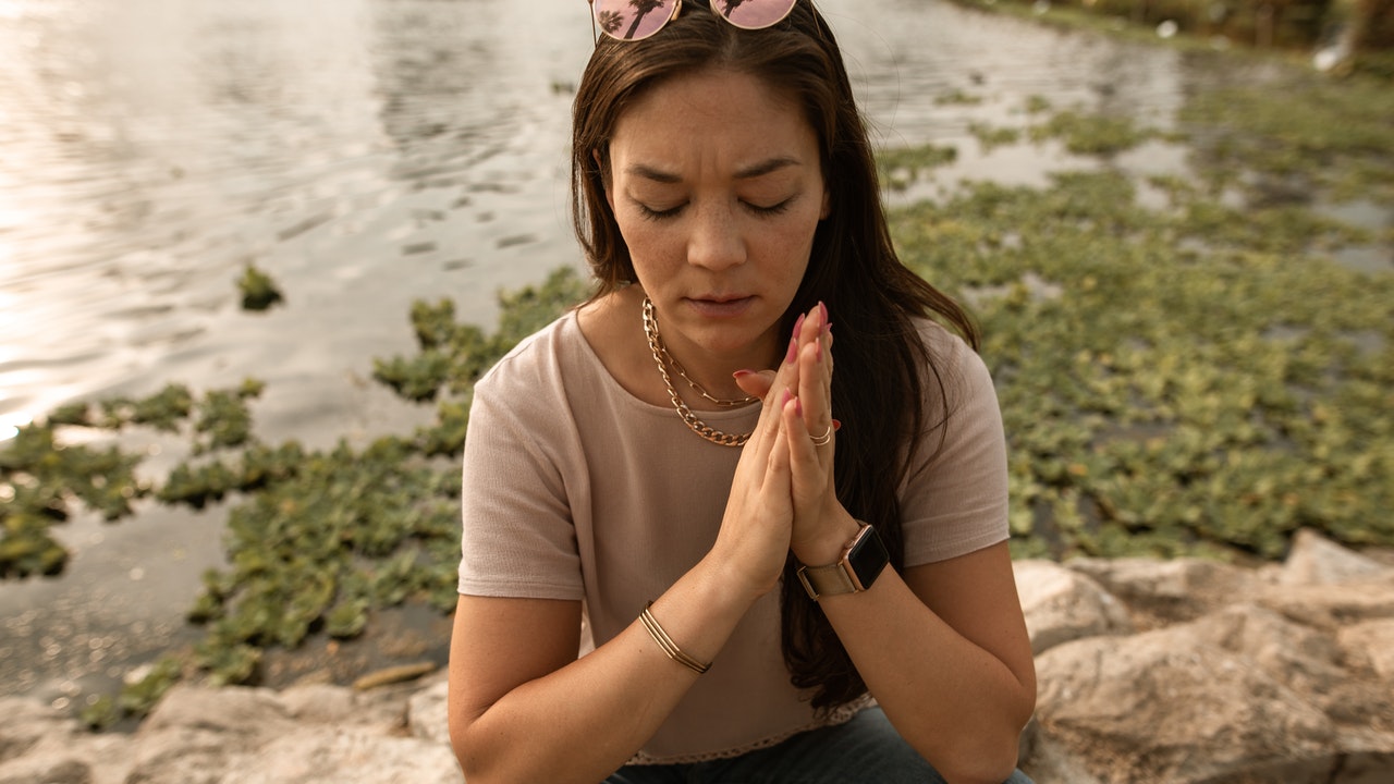 Woman sitting by a Lakeshore with hands clasped and eyes closed in deep prayer.