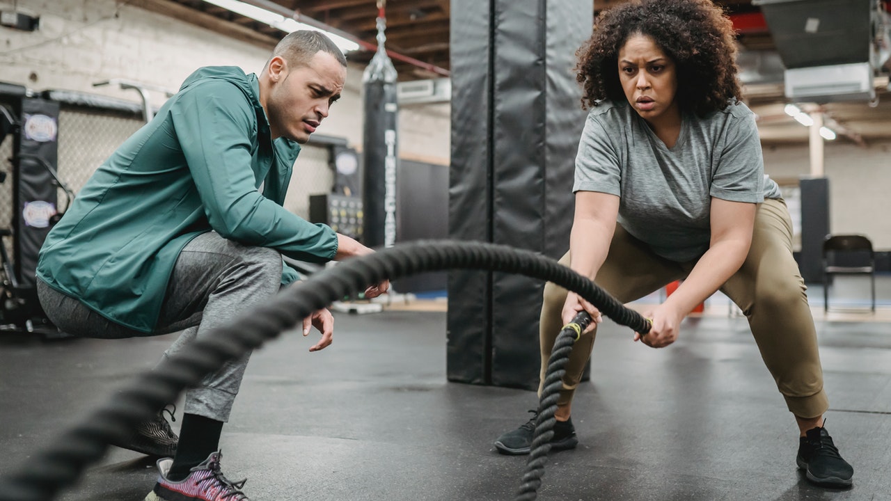 Focused Black woman exercising with battle rope with coach watching.
