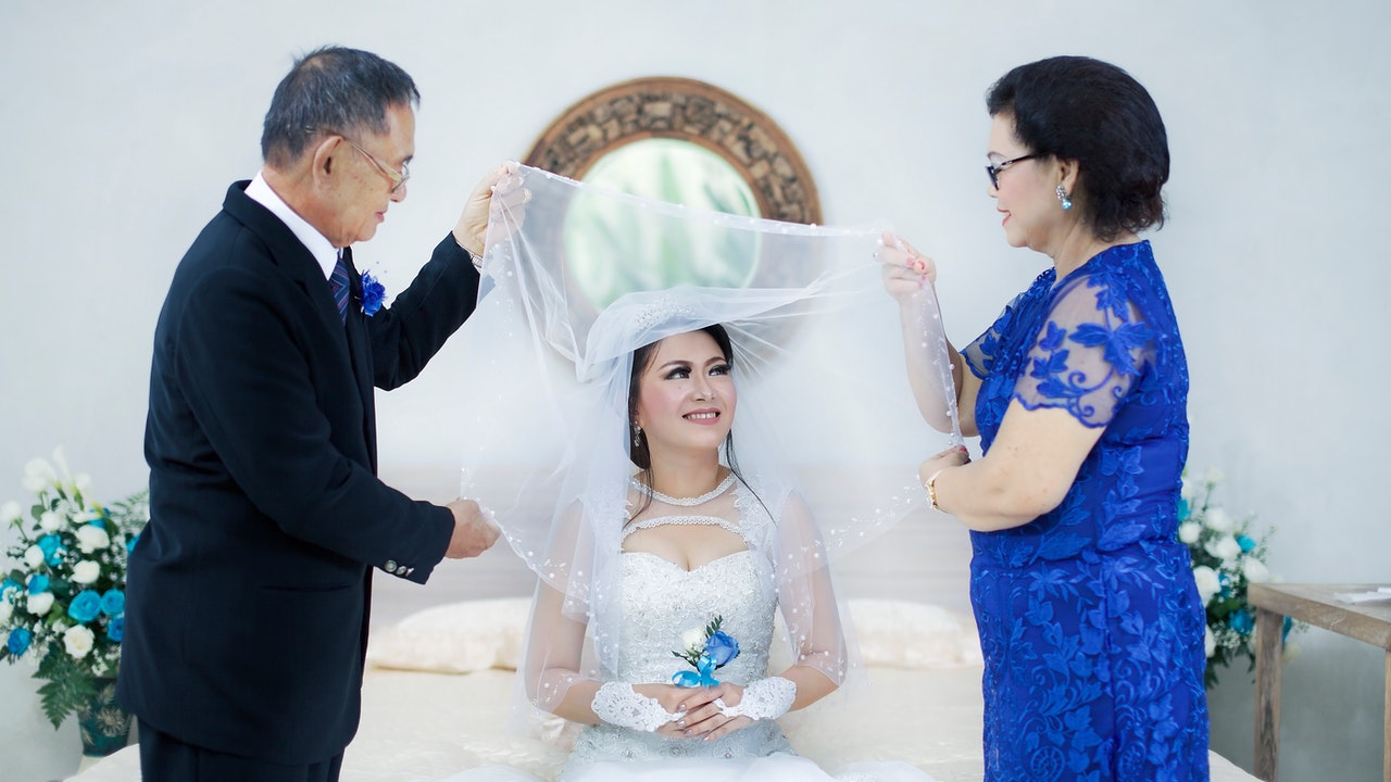A woman in a white wedding gown, dressed and prepared by a man in formal suit on the right and a woman in blue on the left.